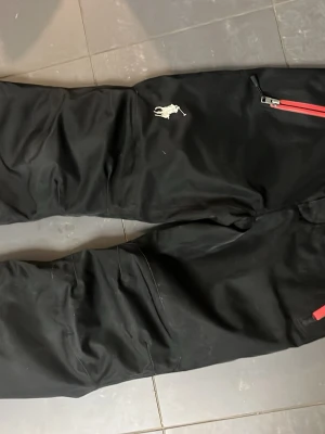 Svarta överdrags byxor  från Polo Ralph Lauren - Snygga svarta cargobyxor från Polo Ralph Lauren med klassisk logga broderad på benet. Byxorna har röda dragkedjor på fickorna och är tillverkade i ett slitstarkt material. Perfekta för en streetwear-look och har flera praktiska fickor. Bra skick inte jätte använda. Priset går att diskutera 🙌 storlek 140