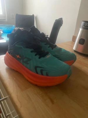 Turkosa och orange sneakers från Hoka - Snygga sneakers från Hoka med turkos ovandel, svarta detaljer och en tjock orange sula. Skorna har svarta skosnören och grafiska mönster på sidan. Perfekta för dig som gillar färgstarka och bekväma sneakers med modern design.