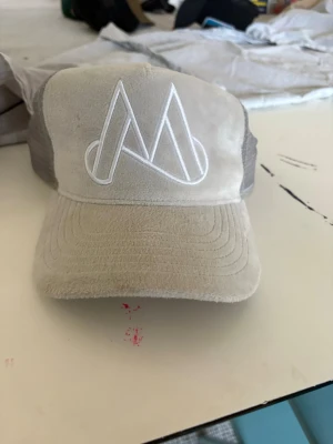 Beige meshkeps med broderad logga - Snygg beige keps med vit broderad logga framtill. Framsidan är i mjuk mockaliknande material och baksidan har grå mesh för extra ventilation. Justerbar snapback för perfekt passform. Perfekt för dig som vill ha en stilren och trendig accessoar.