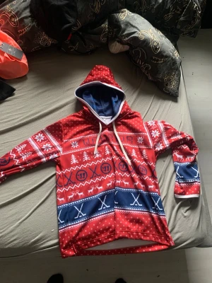 Röd och blå julig hoodie med mönster - Snygg hoodie i rött och blått med julinspirerat mönster, snöflingor, granar och renar. Utsidan är röd med vita och blå detaljer, insidan av huvan är blå. Perfekt för vintern och julen!