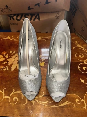 Silvriga glitterpumps från Graceland - Snygga silvriga pumps från Graceland med glitterfinish och öppen tå. Skorna har smal klack och en klassisk peep-toe design. Perfekta för att lyfta vilken outfit som helst med extra bling. Materialet är syntet med glittrig yta.