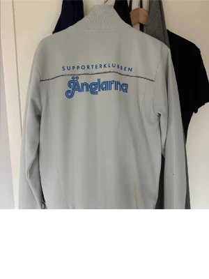 Ljusgrå vindjacka Änglarna - Snygg ljusgrå vindjacka från Änglarna Supporterklubben med blå logga på bröstet och stor text på ryggen. Jackan har hög krage, hel dragkedja framtill och två svarta dragkedjefickor. Perfekt för dig som vill visa ditt supporterskap.