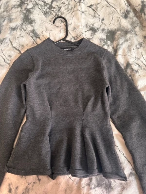 Mörkgrå knitted peplumtröja från Gina Tricot  - Snygg mörkgrå peplumtröja från Gina Tricot med rund halsringning och långa ärmar. Tröjan har en figurnära passform upptill och en utställd nederdel som ger en feminin siluett. Tillverkad i mjukt stickat material som känns bekvämt mot huden.