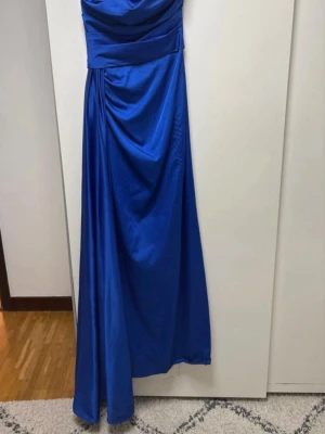 Blå långklänning med drapering - Elegant blå långklänning med draperad överdel och axelbandslös design. Klänningen har en markerad midja och ett vackert fall i kjolen. Perfekt för speciella balen när du vill sticka ut. Materialet känns mjukt och har en lätt glans.               Använd 1 gång på balen för 2 år sedan men förvarad trott mörkt i skydd. Plagget är kemtvättat. Original priset: 3200kr 