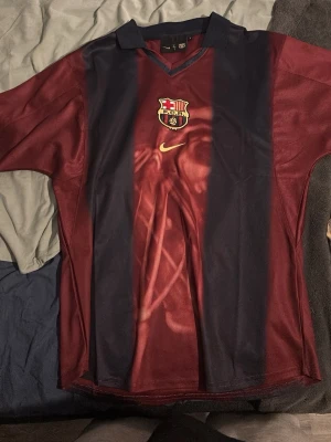 Barcelona x Cactus Jack matchtröja - Unik Barcelona matchtröja i vinrött och marinblått med breda ränder, officiellt klubbmärke och Nike-logga på bröstet. På ryggen står det 'Cactus Jack', nummer 2 och 'BARÇA' i graffiti-stil. Perfekt för dig som älskar fotboll och streetwear.