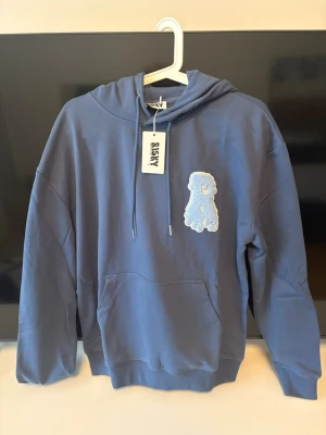 Blå hoodie från Risky med patch - Säljer en blå hoodie från Risky med huva och dragsnören. På bröstet sitter en stor patch av en blå hund och nere på magen finns trycket 'RISKY' i droppande stil. Hoodien har känguruficka och ribbade muddar. Perfekt för dig som gillar streetwear och unika detaljer.