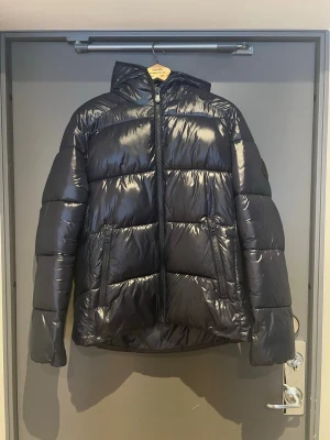 Svart glansig pufferjacka med huva - Säljer en blå pufferjacka med glansig finish och stor huva. Jackan har två dragkedjeförsedda fickor framtill och breda quiltade paneler. Perfekt för kalla dagar och riktigt snygg till streetwear-stilen.