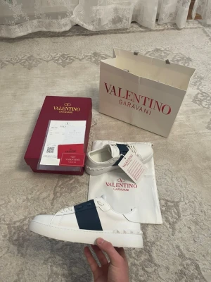Valentino Garavani vita sneakers - Säljer ett par snygga Valentino Garavani sneakers i vitt skinn med marinblå detalj över snörningen. Skorna har rund tå, platt sula och klassisk logga på plösen. Medföljer originalkartong, dustbag och äkthetsbevis. Perfekt för dig som gillar stilrena designer. Lite andvända men inget farligt