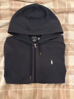 Mörkblå hoodie från Polo Ralph Lauren - Snygg mörkblå hoodie med dragkedja från Polo Ralph Lauren. Klassisk design med vit broderad logga på bröstet, huva med dragsko och två fickor framtill. Perfekt för en avslappnad och stilren look.