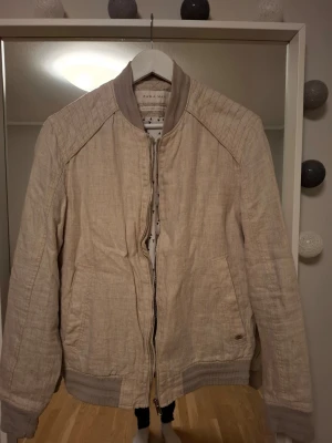 Beige bomberjacka från Zara Man - Stilren beige bomberjacka från Zara Man i ett lätt linneliknande material. Jackan har ribbade muddar vid ärmslut och midja, diskreta sidofickor och dragkedja framtill. Snygga sömdetaljer på axlarna ger extra edge. Perfekt för dig som gillar minimalistisk och clean stil.