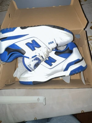 New Balance 550 vita/blå sneakers - Snygga New Balance 550 sneakers i vitt och blått med svarta detaljer. Skorna har en klassisk låg siluett, perforerade paneler och tydliga New Balance-loggor på sidorna. Ovandelen är i läder och textil, och sulan är robust med bra grepp.