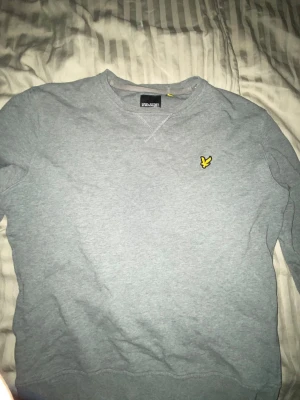 Grå sweatshirt från Lyle & Scott - Snygg grå sweatshirt från Lyle & Scott med klassisk rund halsringning och den ikoniska gula örnloggan broderad på bröstet. Tröjan har ribbade muddar vid ärmslut och nederkant, perfekt för en avslappnad stil.