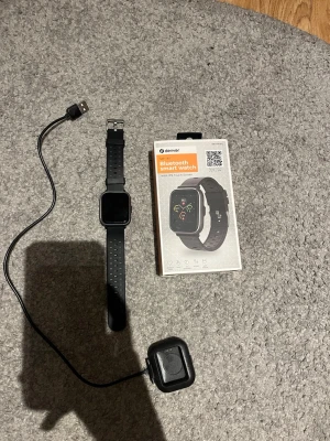 Denver svart smartklocka med laddare - Säljer en svart Denver Bluetooth smartklocka med fyrkantig display och silikonarmband. Klockan har pulssensor och levereras med originalförpackning och laddare. Perfekt för dig som vill ha koll på notiser, träning och hälsa direkt på handleden.