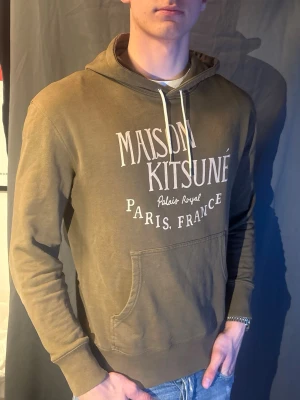 Maison Kitsuné hoodie olivgrön - Snygg olivgrön hoodie från Maison Kitsuné med vit text på bröstet: 'Maison Kitsuné Palais Royal Paris, France'. Modellen är 190cm och väger 80kg. Skriv om du har frågor 🥰