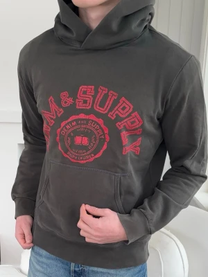 Mörkgrå hoodie från Ralph Lauren denim and supply  - Snygg mörkgrå hoodie från Ralph Lauren med röd text och logga på bröstet. Klassisk känguruficka framtill och stor huva. Tillverkad i mjukt bomullsmaterial som känns skönt mot huden. Perfekt för en avslappnad och cool stil.