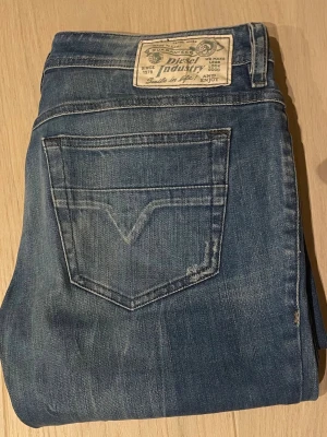 Blå jeans från Diesel - Säljer ett par klassiska blå jeans från Diesel med raka ben och snygga slitningar. Jeansen har två bakfickor med V-formad söm och en patch med Diesel Industry-logga i midjan. Tillverkade i slitstark denim som ger en cool och avslappnad look.