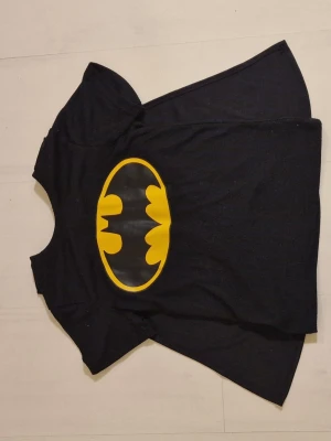 Batman t-shirt med cape - Cool svart Batman t-shirt med gul Batman-logga på bröstet och kardbordbands fast cape baktill. Perfekt för maskerad eller cosplay. Tillverkad i mjukt tyg med korta ärmar och rund hals. Snygg och bekväm för alla superhjältefans!