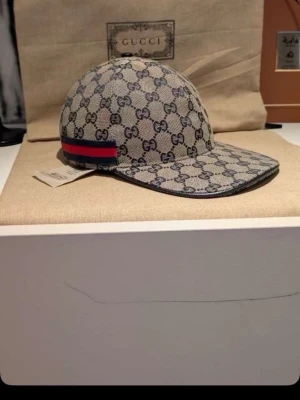 Gucci keps med GG-mönster - Säljer en snygg keps från Gucci med klassiskt GG-monogram i beige och mörkblått. Kepsen har en böjd skärm och en röd-blå rand på sidan som ger extra lyxig känsla. Perfekt accessoar för dig som vill sticka ut med stilren design och exklusivt märke.
