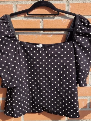 Svart prickig topp från H&M - Snygg svart topp från H&M med vita prickar och puffiga korta ärmar. Toppen har en fyrkantig ringning och är gjord i mjuk viskos o polymid blandning.Perfekt för dig som gillar trendiga detaljer och klassiskt mönster.Med puffärmar sitter den väldigt fint på och har stretch vid axlarna så den kan bäras både vanligt och off-shoulder. Fint skick!"