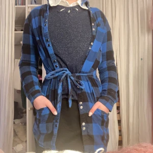 Blå och svart rutig kofta med knytband - Snygg lång kofta i blått och svart rutigt mönster. Koftan har knappar framtill, ett matchande knytband i midjan och långa ärmar. Perfekt för lager på lager och ger en cool, avslappnad look.