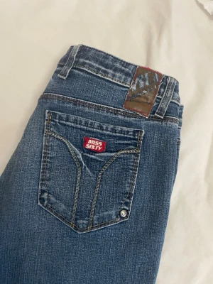 Blå Miss Sixty bootcut jeans - Snygga blå jeans från Miss Sixty med bootcut passform och klassisk femficksdesign. Jeansen har kontrastsömmar, läderpatch bak i midjan och en röd Miss Sixty-logga på bakfickan. Tillverkade i stretchigt denim för skön passform. Utsydda där nere till bootcut.
