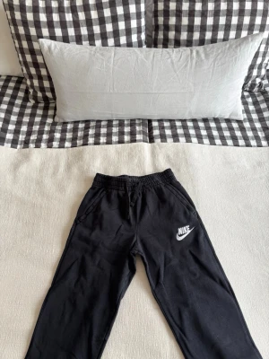 Svarta mjukisbyxor från Nike - Säljer ett par svarta mjukisbyxor från Nike med klassisk vit logga på vänster ben. Byxorna har resår i midjan med dragsko och sidofickor. Perfekta för chill dagar eller träning. Materialet känns mjukt och bekvämt mot huden.
