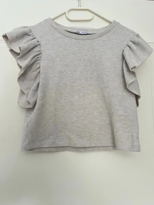 Beige topp med volangärmar Zara - Supersöt beige topp med volangdetaljer på ärmarna och rund halsringning. Tillverkad i mjukt material. Använd någon få gång! Lite kortare i modellen