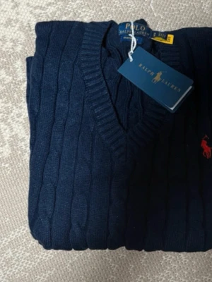 Mörkblå stickad tröja Polo Ralph Lauren - Klassisk mörkblå stickad tröja från Polo Ralph Lauren med kabelstickat mönster och V-ringning. Tröjan har den ikoniska röda logotypen broderad på bröstet och ribbade muddar. Perfekt för en stilren och tidlös look.