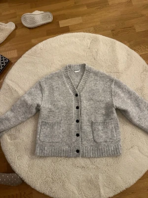 Ljusgrå kofta från Weekday - Mysig ljusgrå kofta från Weekday med v-ringning, knappar framtill och två stora fickor. Koftan är i storlek S och passar perfekt till allt! Nästan aldrig använd