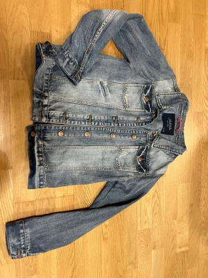 Blå jeansjacka  - Klassisk blå jeansjacka  med slitna detaljer och kopparfärgade knappar. Två bröstfickor med lock och knapp, samt raka sömmar och normal passform. Perfekt till en avslappnad streetstyle-look.