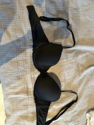 Svart push-up bh från Emilia - Snygg svart push-up bh från Emilia med vadderade kupor och spetsdetaljer på sidorna. Justerbara axelband och klassisk hakknäppning bak. Perfekt för dig som vill ha extra lyft och en stilren look.