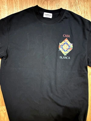 Svart t-shirt från Casablanca - Svart t-shirt från Casablanca med färgglatt tryck av sol, fåglar och flygplan på ryggen samt mindre tryck på bröstet. Rund halsringning och avslappnad passform. Perfekt för dig som gillar unika prints och streetwear vibes.