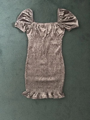 Grå sammetsklänning med puffärm - Supersöt kort grå klänning i mjuk sammet med puffiga kortärmar och smockad kropp. Klänningen har volangkant nertill som ger en trendig vibe. Perfekt för dig som vill sticka ut med en unik och bekväm stil. Använt endast en gång, inga slitage!