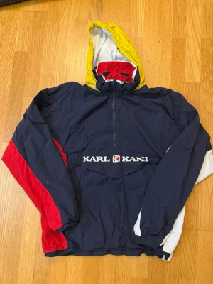 Flerfärgad vindjacka från Karl Kani - Retro vindjacka från Karl Kani i marinblått med röda, vita och gula detaljer. Jackan har huva, halv dragkedja framtill och stor ficka med broderad logga på magen. Oversized passform och elastiska muddar. Perfekt för streetwear-stil.