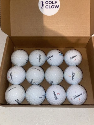 Titleist mix 12-pack - Säljer ett set med tolv vita golfbollar från Titleist, levererade i en brun kartong med Golf Glow-klistermärke. Bollarna har klassisk dimple-design och märkning med siffror och logga. Perfekt för dig som vill fylla på golfutrustningen.