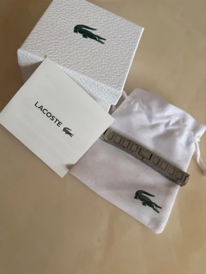 Silverfärgat armband från Lacoste - Snyggt oanvänd armband från Lacoste i silverfärgad metall med stilrena länkar och diskret Lacoste-logga. Kommer med original ask och vit tygpåse med den klassiska gröna krokodilen. Perfekt accessoar för dig som gillar en clean och modern look. Den är cirka 20 cm lång men kan enkelt ändras jag kan även hjälpa till om det behövs😃😃