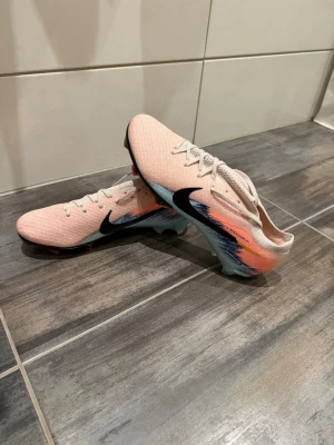 Nike Mercurial rosa fotbollsskor - Snygga Nike Mercurial fotbollsskor i ljusrosa med svart swoosh och färgstänk i blått och orange på sidan. Skorna har låg profil, snörning och en tunn ovandel i syntetmaterial för bra bollkontroll. Perfekta för dig som vill sticka ut på planen.