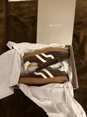 Bruna sneakers från Gant i mocka - Snygga bruna sneakers från Gant i mocka med vita detaljer och gummisula. Klassisk låg modell med svarta skosnören och retroinspirerad design. Perfekta för dig som gillar stilrena och tidlösa sneakers med en sportig touch. Helt nya