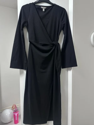 Svart omlottklänning med knyt - Svart långärmad omlottklänning med knyt i midjan. Klänningen har en v-ringad halsringning och är i ett mjukt, lätt material som ger en snygg siluett. Perfekt för dig som gillar stilrena och enkla plagg med en twist.