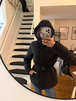 Svart pufferjacka med bälte - Säljer en svart pufferjacka med huva och bälte i midjan. Jackan har en stilren design med quiltade partier och är perfekt för kalla vinterdagar. Passar dig som vill ha en varm och trendig jacka med snygg passform.