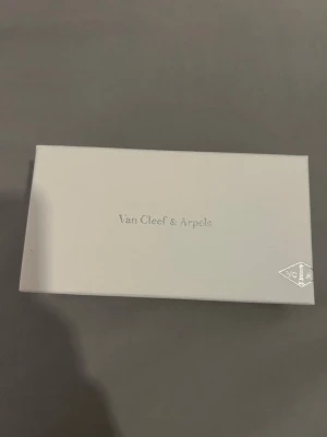 Smyckesask Van Cleef & Arpels - Elegant smyckesask från Van Cleef & Arpels i ljusbeige och ljusgrön färg. Asken har en stilren design med logotypen tryckt på både utsidan och insidan av locket. Perfekt för att förvara exklusiva smycken.