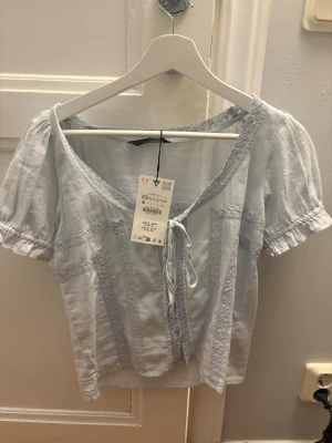 Ljusblå blus med knyt och spetsdetaljer - Supersöt ljusblå blus från Zara  med korta puffärmar, knyt framtill och fina spetsdetaljer längs kanterna. Croppad modell som ger en fräsch och somrig vibe. Perfekt att matcha med jeans eller kjol för en trendig look.