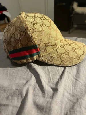 Beige Gucci keps med GG-mönster - Säljer en beige keps från Gucci med det klassiska GG-monogrammet över hela kepsen. På sidan finns de ikoniska gröna och röda ränderna. Kepsen har böjd skärm och är tillverkad i ett slitstarkt tygmaterial. Perfekt för dig som vill ha en stilren och lyxig accessoar.