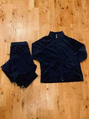 Mörkblå velour loungewear-set - Mysigt loungewear-set i mörkblå velour med dragkedja på tröjan och resår i midjan på byxorna. Setet har en sportig look med hög krage och raka byxben, perfekt för chill dagar hemma. Materialet är mjukt och glansigt för extra komfort. Byxbenen har söta rosett detaljer som man kan knyta för tajtare byxkant.