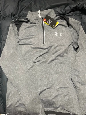 Grå träningströja från Under Armour - Snygg grå långärmad träningströja från Under Armour med halv dragkedja och hög krage. Tillverkad i ett funktionsmaterial som andas, perfekt för löpning eller gym. Diskret logga på bröstet och svarta detaljer vid axlarna.