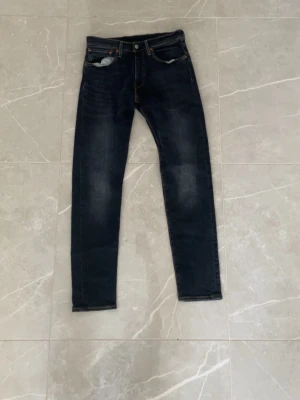 Mörkblå raka jeans med kontrastsömmar - Säljer ett par mörkblå jeans med raka ben och snygga kontrastsömmar. Klassisk femficksmodell med knappgylf och detaljer i kopparfärg. Perfekta till en avslappnad stil och passar till det mesta. Materialet är denim i bomull.