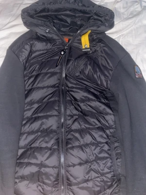 Svart pufferjacka från Parajumpers - Säljer en svart pufferjacka från Parajumpers med huva och dragkedja. Jackan har quiltad front, släta ärmar och en gul detalj vid halsen. Märkespatch på ärmen och innerfoder i fleece. Perfekt för kalla dagar och riktigt snygg streetstil.