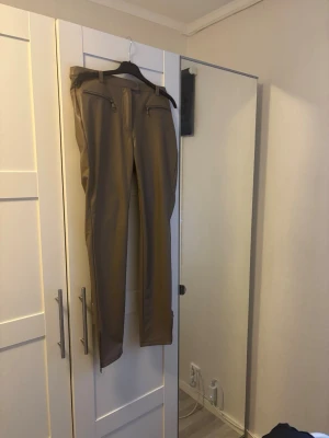 Beige byxor med dragkedjedetaljer - Snygga beige byxor från Only i skinnimitation med raka ben och dragkedjor vid fickorna framtill. Byxorna har en klassisk passform och stängs med knapp och dragkedja. Perfekta för dig som vill ha en stilren och enkel look. OBS! Tight modell och liten i stk 40. Inte användna. 