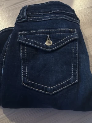 Mörkblå jeans med kontrastsömmar - Snygga mörkblå jeans med vita kontrastsömmar och ficklock med knapp bak. Klassisk femficksmodell och normal midja. Jeansen har en stilren look och passar perfekt till en avslappnad outfit. Materialet är troligtvis bomull med stretch för extra komfort.