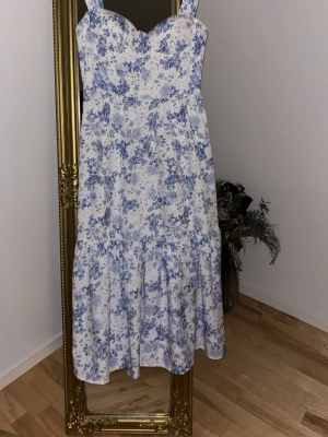 Blommig långklänning från Covenia - Romantisk långklänning från Covenia i vit bomull med blått blommönster. Klänningen har breda axelband, markerad byst och volang nedtill för en feminin siluett. Perfekt för sommarens alla tillfällen.